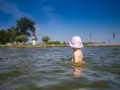 Kleines Kind mit rosa Hut planscht im Wasser am Stettiner Haff Strand. Im Hintergrund Bäume und ein Leuchtturm.