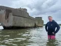 Mann steht im Wasser neben einem verwitterten Betonschiff in Wismar.