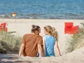 Ein Paar sitzt im Sand am Trassenheide Strand, blickt sich an. Im Hintergrund das Meer und rote Strandkörbe.