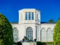 Die Orangerie Putbus, ein strahlend weißes Gebäude mit Skulpturen und großen Fenstern, wird von üppigem Grün eingerahmt.