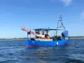 Blaues Fischerboot "RERIK" mit roten Markierungen auf dem Wasser. Ein Mann steht an Deck, rote Fahnen ragen empor.