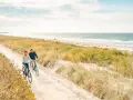 Ein Paar genießt seine baltischen Abenteuer auf Fahrrädern entlang eines sandigen Weges am Meer, umgeben von Dünen und Gräsern.