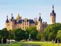 Das Schweriner Schloss, ein Märchenschloss mit goldenen Kuppeln und Türmen, thront majestätisch über einem grünen Park mit Statuen und einem Teich.
