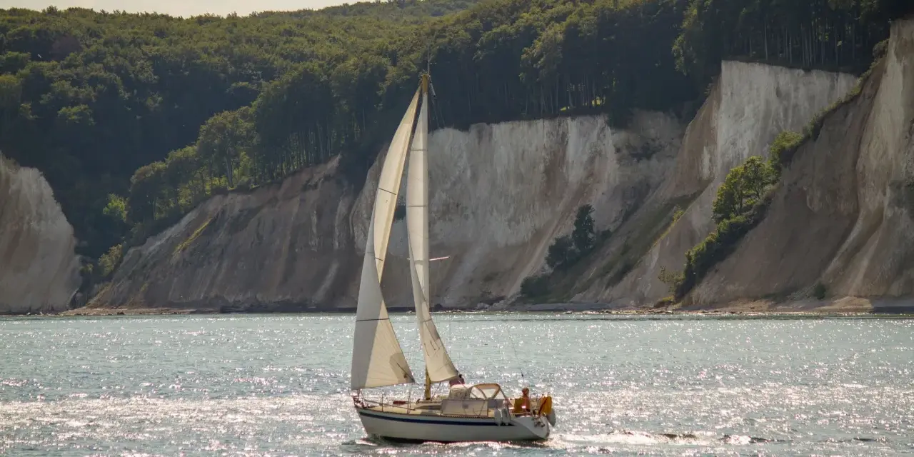 Segelboot gleitet über glitzerndes Wasser vor Kreidefelsen und grünen Wäldern. Ein Tag zum Ostsee segeln.