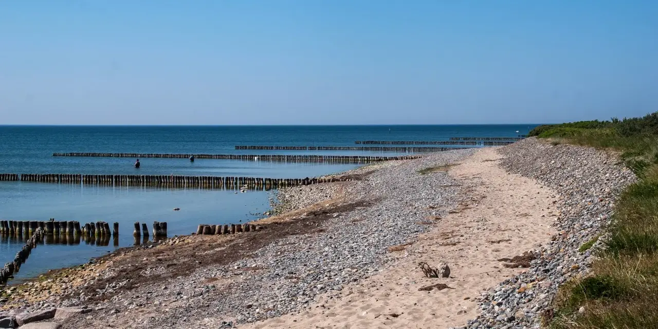 Ein ruhiger Tag am Dranske Strand. Holzpfähle ragen aus dem ruhigen Meer, das sich unter einem klaren blauen Himmel erstreckt.