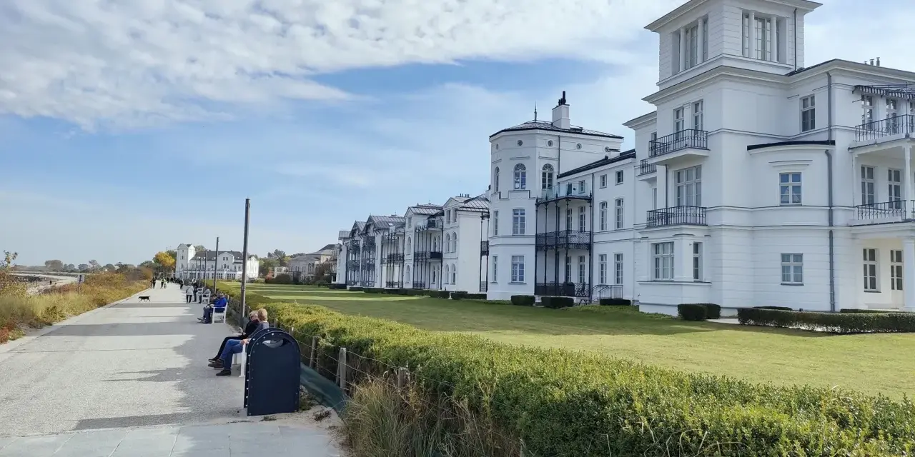 Weiße Villen säumen die Heiligendamm Promenade. Menschen spazieren und genießen die Aussicht auf das Meer.