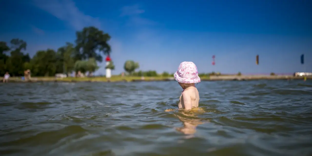Kleines Kind mit rosa Hut planscht im Wasser am Stettiner Haff Strand. Im Hintergrund Bäume und ein Leuchtturm.