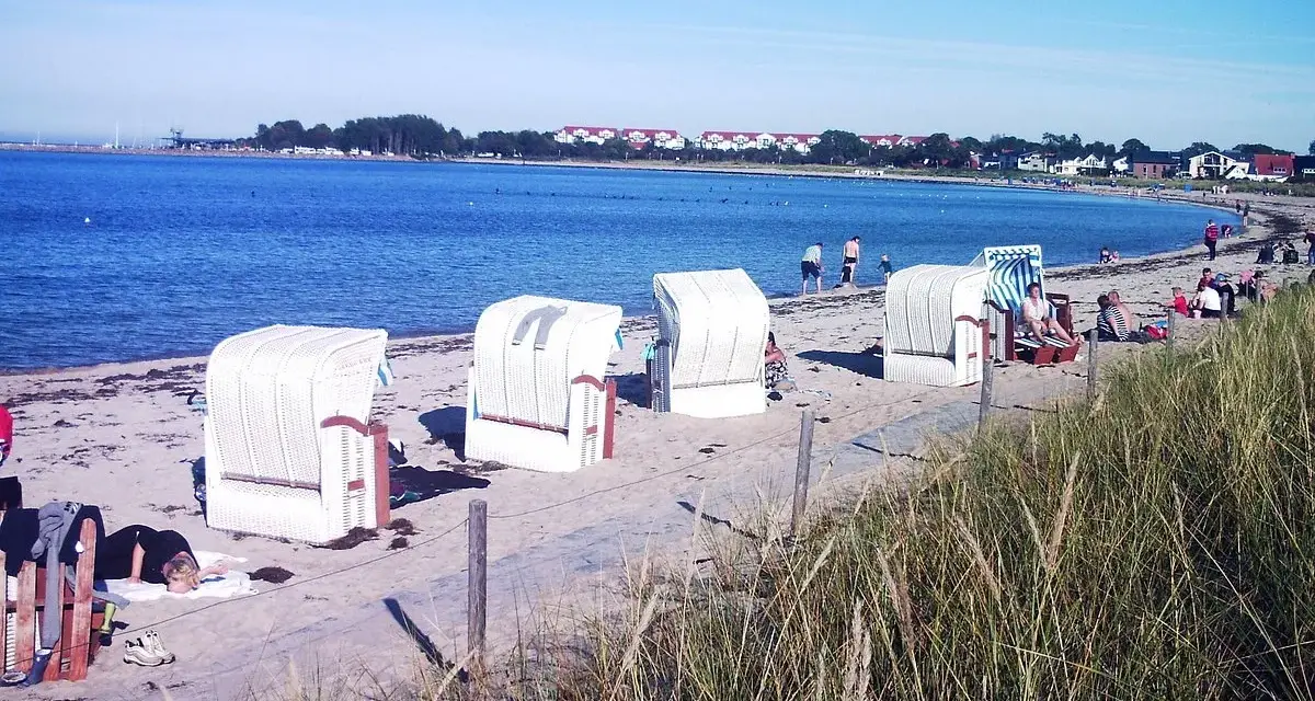 Strandkörbe am Ostseestrand, im Hintergrund der glowe hafen mit Häusern und Bäumen.