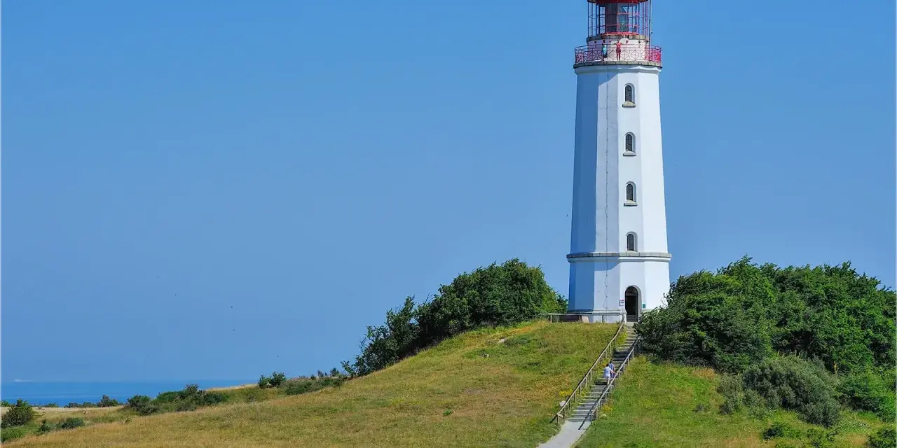 Der Leuchtturm Dornbusch ragt auf einer grünen Anhöhe empor, mit dem blauen Meer im Hintergrund. Eine Treppe führt zum Eingang.