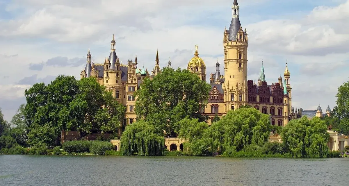 Ein märchenhaftes Schloss mit vielen Türmen und goldenen Kuppeln, umgeben von grünen Bäumen, spiegelt sich im Wasser. Ein gut gremmelin-artiger Anblick.