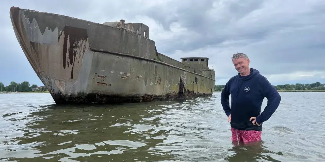 Mann steht im Wasser neben einem verwitterten Betonschiff in Wismar.