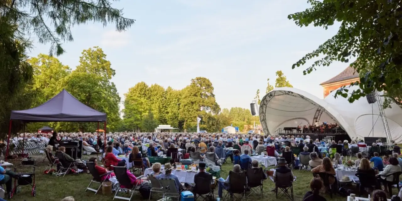 Großes Publikum bei einem Open-Air-Konzert im Rahmen eines **Festivals Mecklenburg-Vorpommern**. Eine Bühne mit Orchester, viele Sitzende auf Rasenflächen.