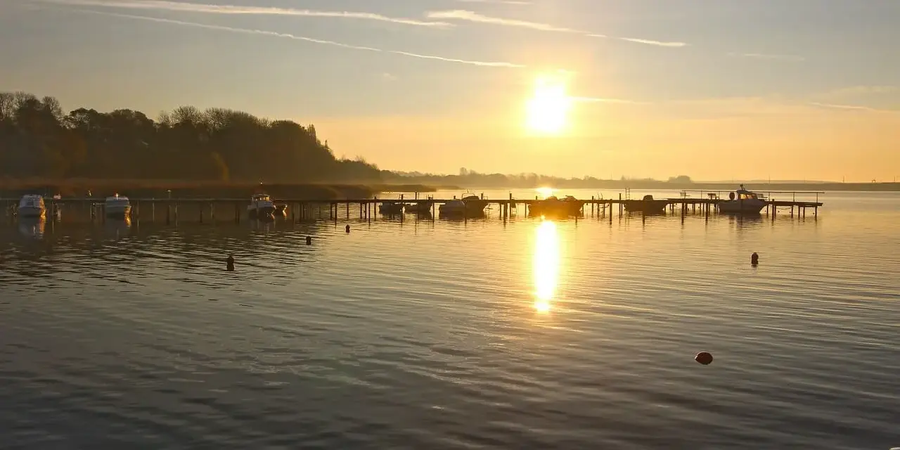 Sonnenaufgang am **Stralsund Strand**. Boote liegen am Steg, die Sonne spiegelt sich im ruhigen Wasser.