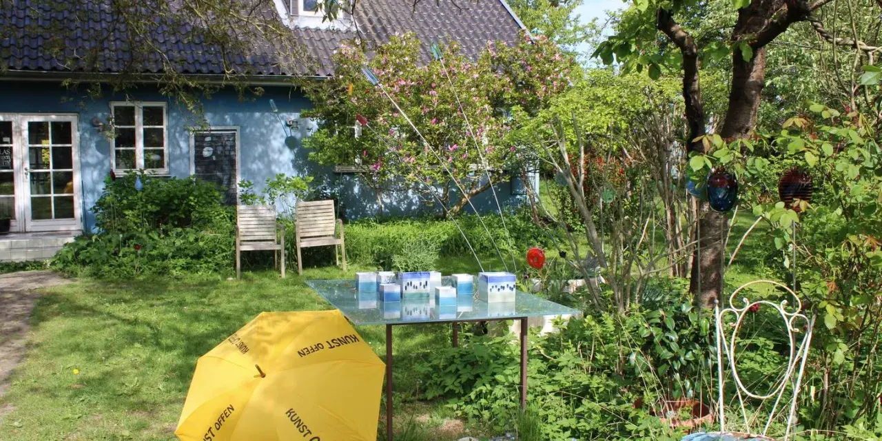 Ein Garten mit blauem Haus, gelbem Regenschirm (Kunst Offen) und Glaskunst auf einem Tisch. Pfingsten 2026 MV.