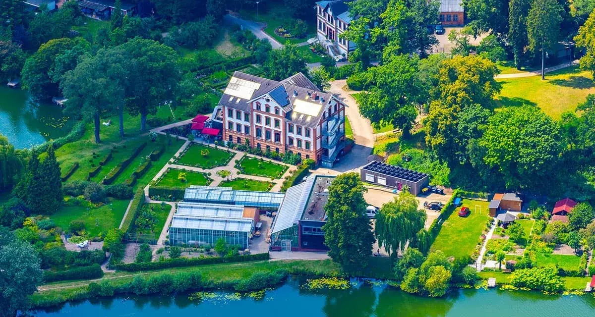 Luftaufnahme des schaugarten am tiefwarensee mit Gewächshäusern, einem Hauptgebäude und gepflegten Gärten am Ufer.