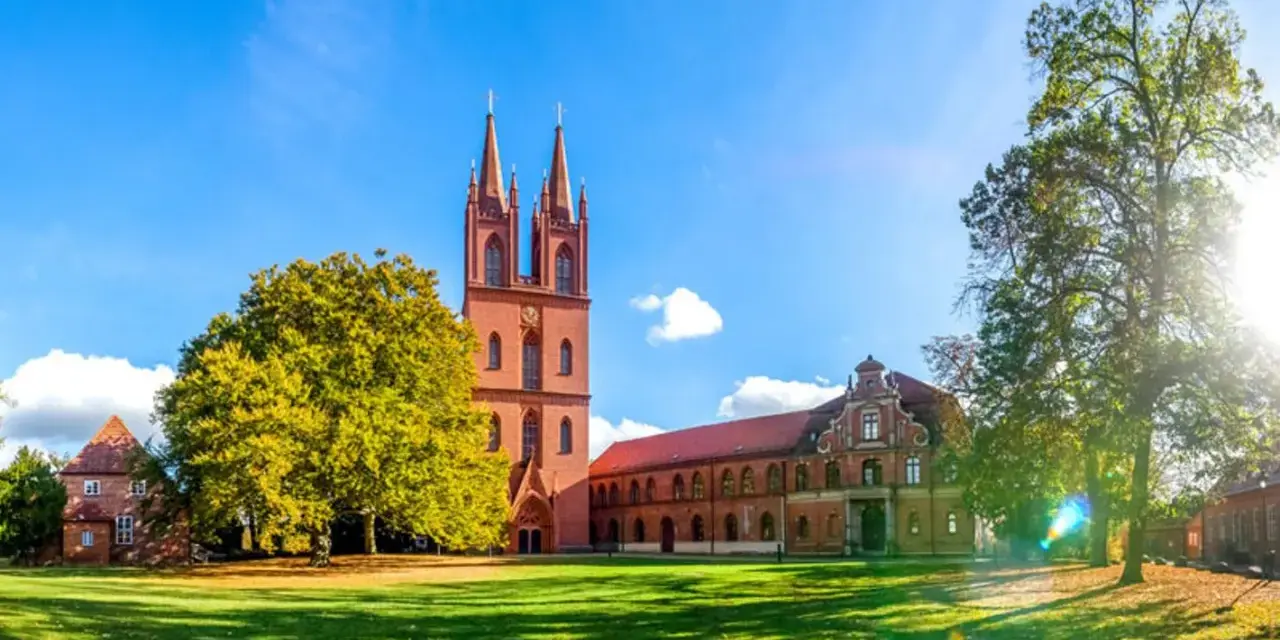 Das Kloster Dobbertin erstrahlt im Sonnenschein. Ein imposanter Kirchturm mit zwei Spitzen ragt empor, umgeben von grünen Rasenflächen und alten Bäumen.