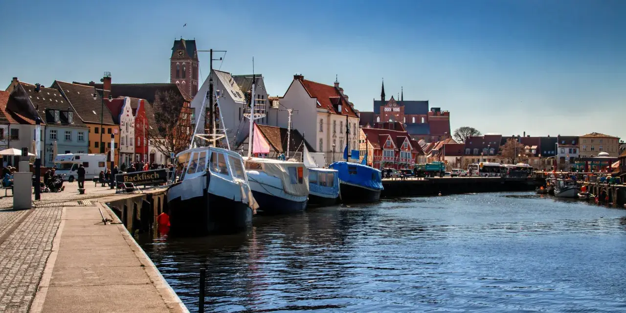 Boote liegen im **Wismar Hafen** vor bunten Häusern. Ein Schild wirbt für "Backfisch".