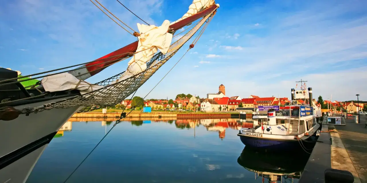 Ein Segelschiff im Vordergrund, im Hintergrund die Stadt Wolgast mit roten Dächern und einem Turm. Ein kleines Boot mit dem Namen "Wolgast" liegt am Kai.