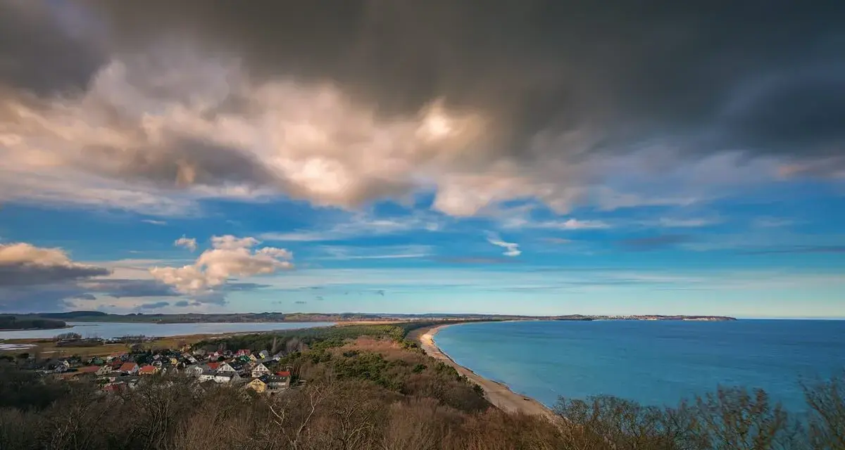 Blick vom Lotsenturm Thiessow über die Küste mit Dorf, Strand und Meer unter dramatischem Himmel.