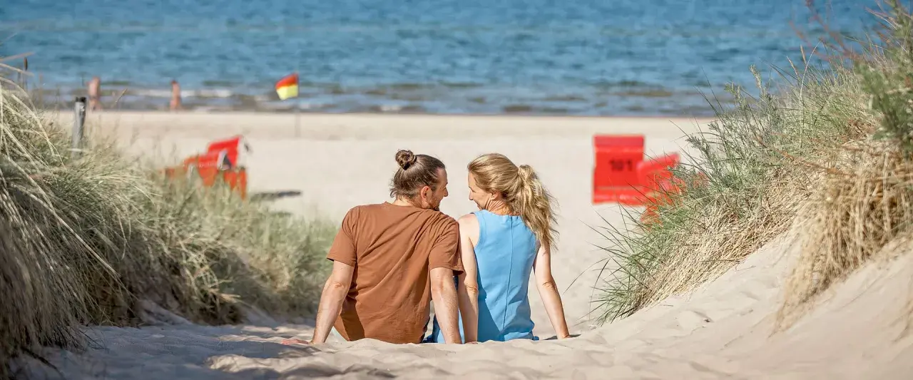 Ein Paar sitzt im Sand am Trassenheide Strand, blickt sich an. Im Hintergrund das Meer und rote Strandkörbe.