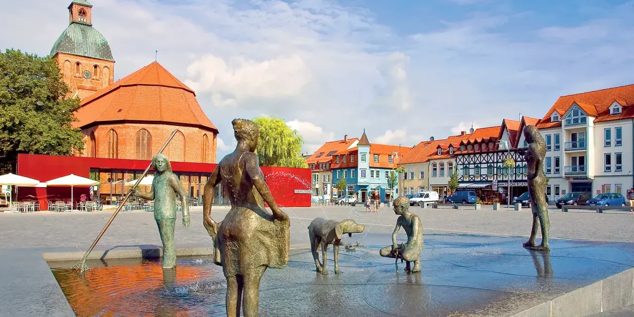 Bronzefiguren am Brunnen in Ribnitz-Damgarten, mit Kirche und Fachwerkhäusern im Hintergrund. Ein Ort, der an Bernstein erinnert.