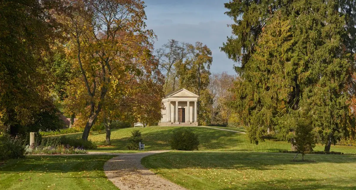 Herbstlicher Spaziergang im Schlossgarten Neustrelitz. Ein Weg führt zu einem antiken Mausoleum, umgeben von bunten Bäumen.