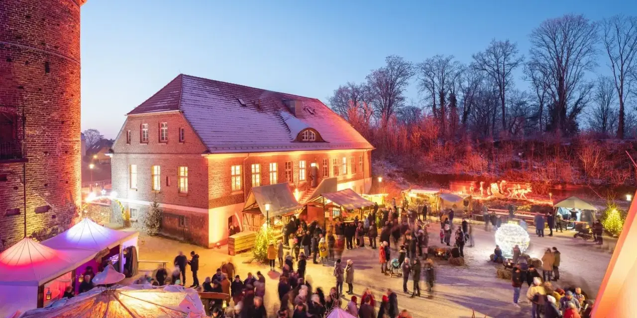 Belebter weihnachtsmarkt plau am see vor historischer Kulisse mit beleuchteten Buden und vielen Besuchern.