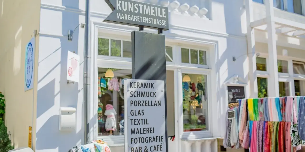 Schild an der Kunstmeile Binz: Keramik, Schmuck, Porzellan, Glas, Textil, Malerei, Fotografie, Bar & Café. Bunte Schals hängen davor.