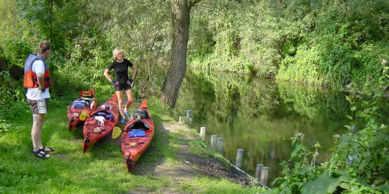 Am Ufer der Kanustation Mirow stehen drei rote Kajaks bereit. Ein Mann mit Schwimmweste und eine Frau in Sportkleidung warten auf ihre Paddeltour.