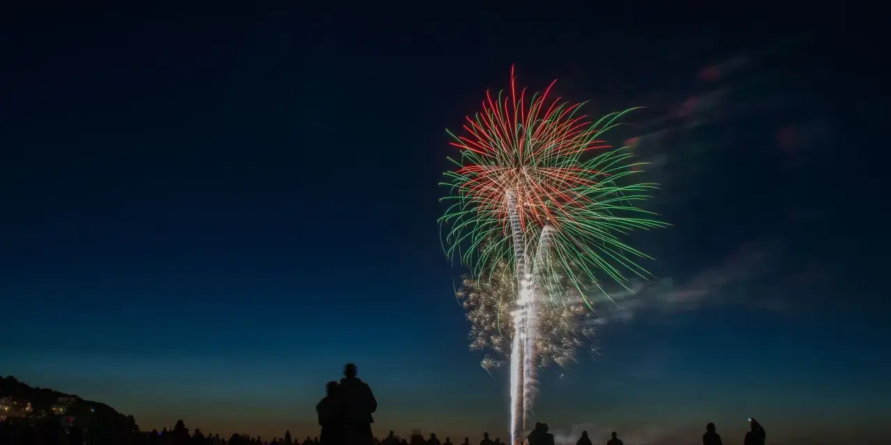 Silhouetten von Menschen am Strand beobachten ein beeindruckendes Feuerwerk über dem Meer. Ein leuchtendes Spektakel in Rot und Grün erhellt den Nachthimmel. Feuerwerk Usedom.