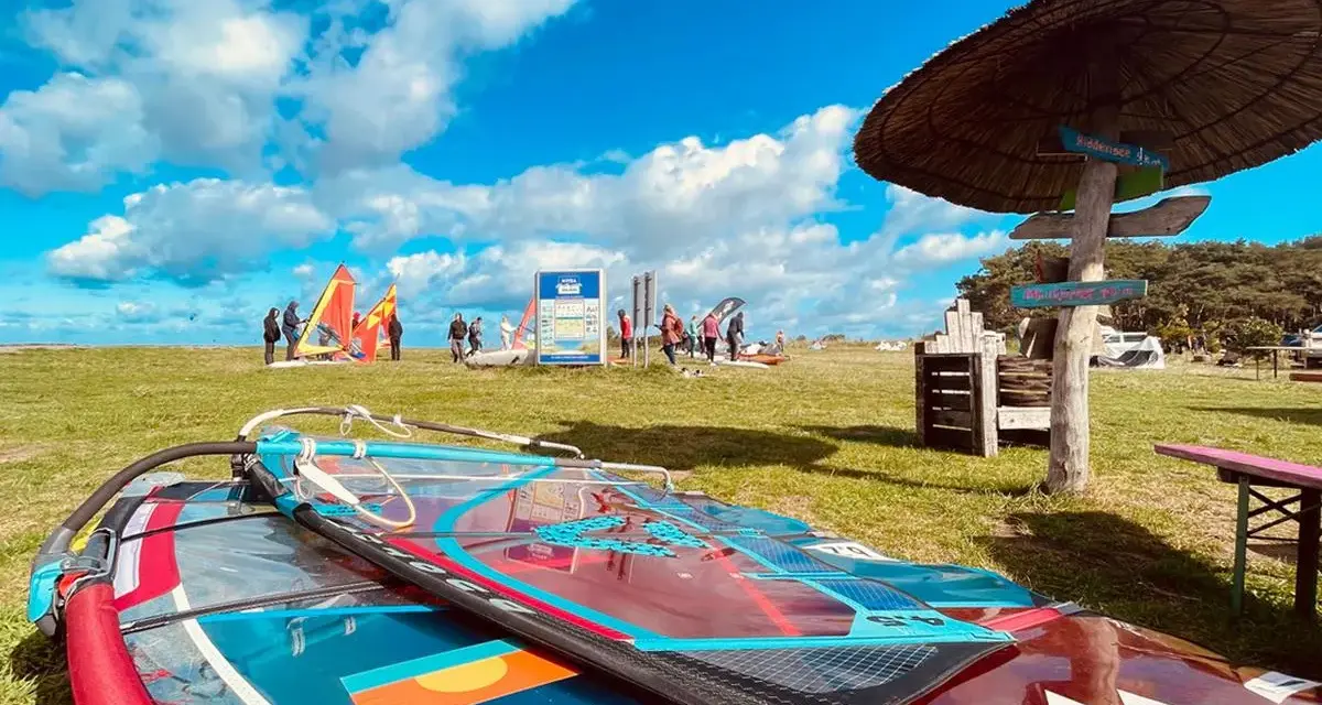 Windsurfausrüstung liegt am ummanz strand. Im Hintergrund bunte Segel und Menschen, die sich auf das Wasser vorbereiten.