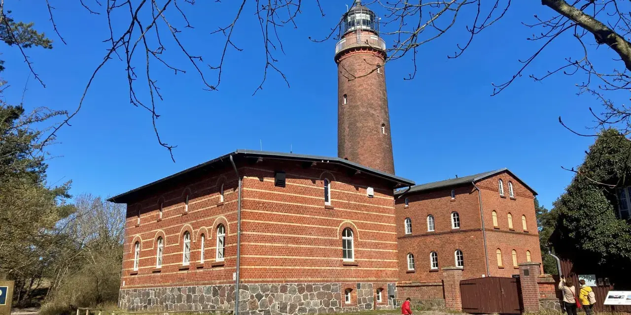 Der Leuchtturm Prerow ragt imposant in den blauen Himmel. Ziegelbauten mit Rundbogenfenstern säumen seinen Fuß.