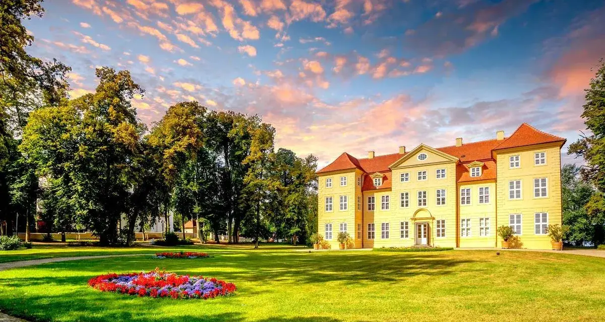 Gelbes Schloss mit roten Dächern, umgeben von grünen Bäumen und Blumenbeeten. Ein malerischer Anblick auf der Liebesinsel Mirow.