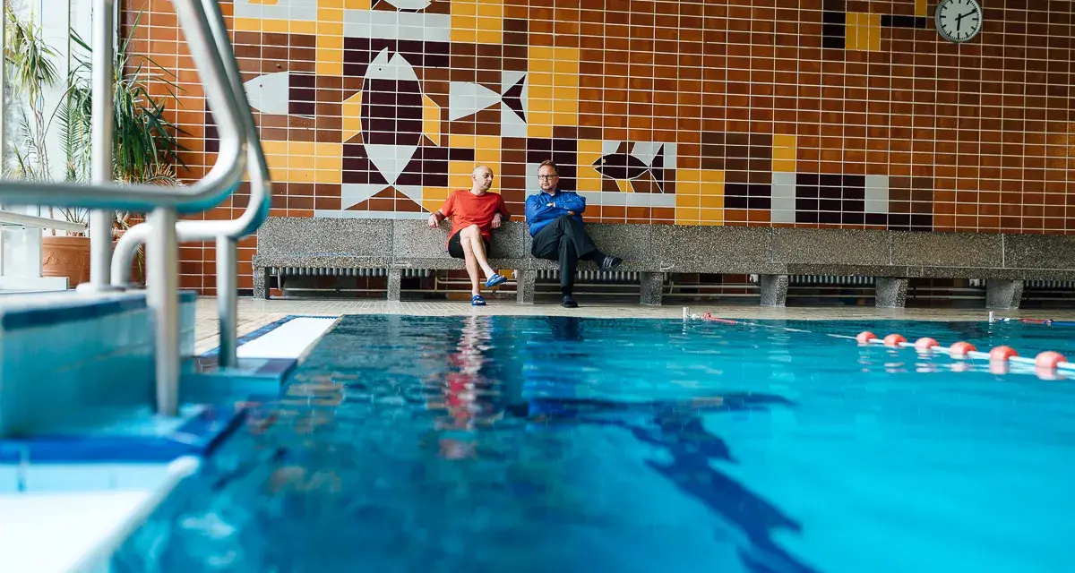 Zwei Männer sitzen in der Schwimmhalle Anklam auf einer Bank vor einer bunten Kachelwand mit Fischmotiven.