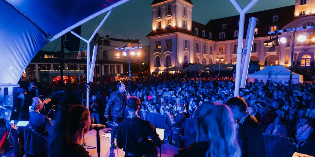 Konzert mit Sänger und Band auf der Bühne, beleuchtet von blauen Lichtern. Im Hintergrund die beleuchtete Architektur und eine Menschenmenge bei Veranstaltungen am Binz Kurplatz.