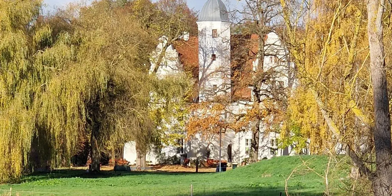 Herbstliche Farben umgeben das Wasserschloss Quilow. Ein Teich spiegelt die Bäume und das weiße Gebäude mit seinem markanten Turm.