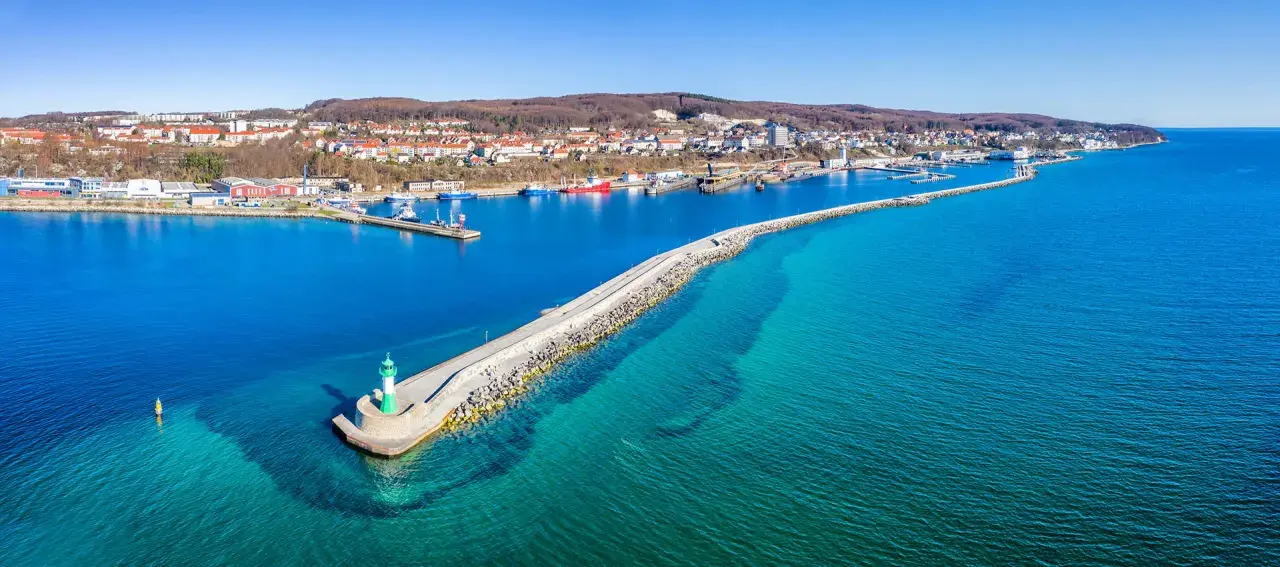 Leuchtturm am Ende einer Mole, die in das klare Wasser vor Sassnitz führt. Boote und die Stadt im Hintergrund.