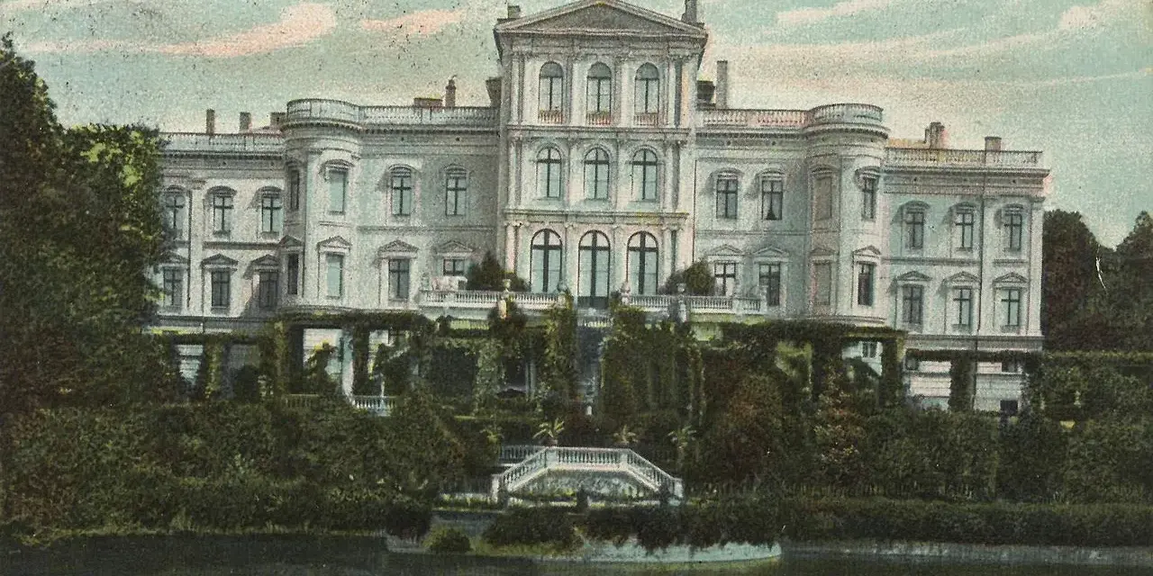 Das Schloss Putbus, von der Rückseite betrachtet, thront majestätisch über einem üppigen Garten und einem ruhigen Gewässer.