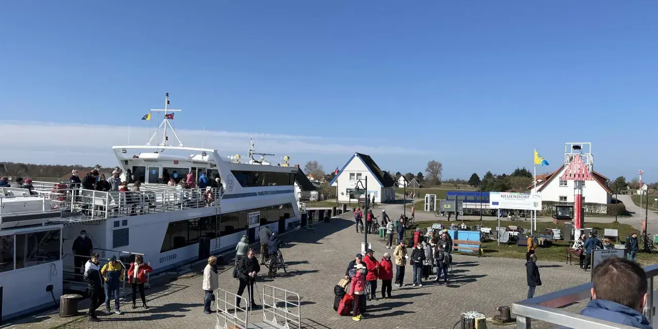 Menschen warten am Kai in Neuendorf auf Hiddensee auf die Fähre. Ein großes Ausflugsschiff liegt bereit.