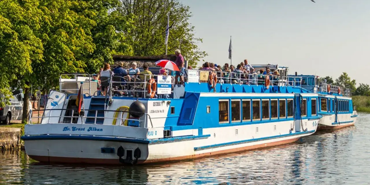 Fahrgastschifffahrt Wichmann: Zwei blau-weiße Schiffe mit Passagieren auf dem Wasser, umgeben von grünen Bäumen und klarem Himmel.