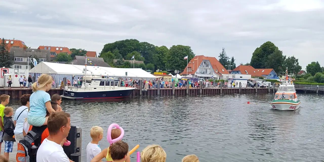 Menschen genießen das bunte Treiben beim Rock am Hafen. Boote liegen im Wasser, ein Rettungsboot fährt vorbei.