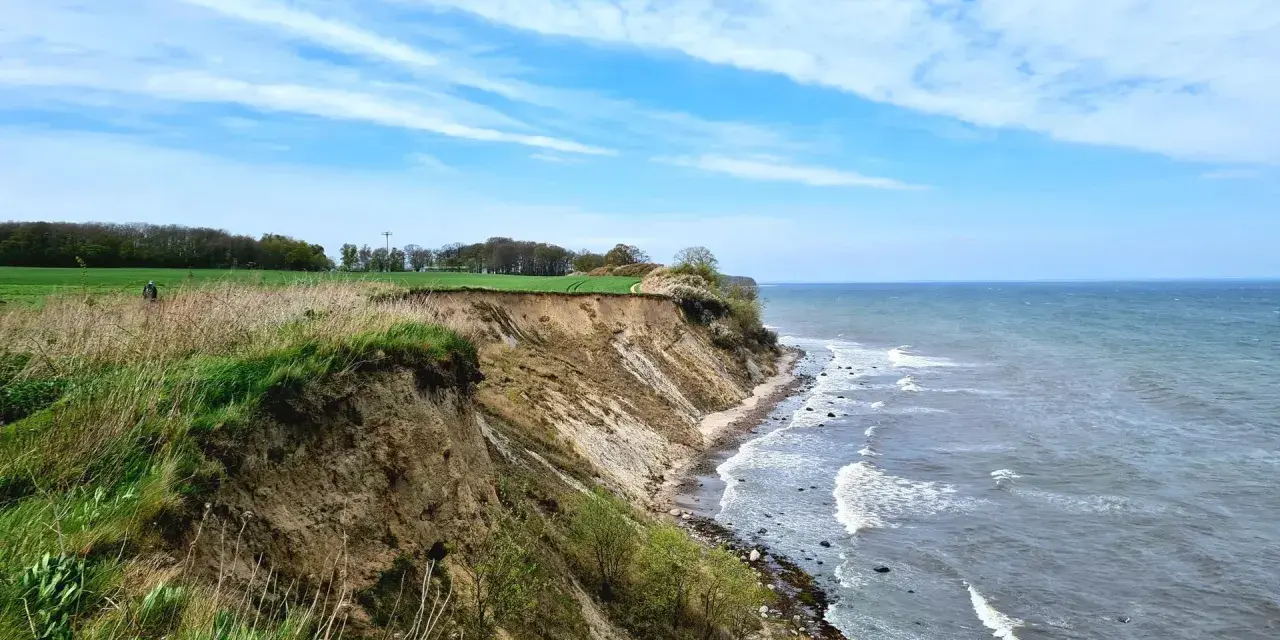 Steilküste mit grasbewachsenem Abhang, der zum steinbeck Strand und dem Meer abfällt. Wolken ziehen über den blauen Himmel.