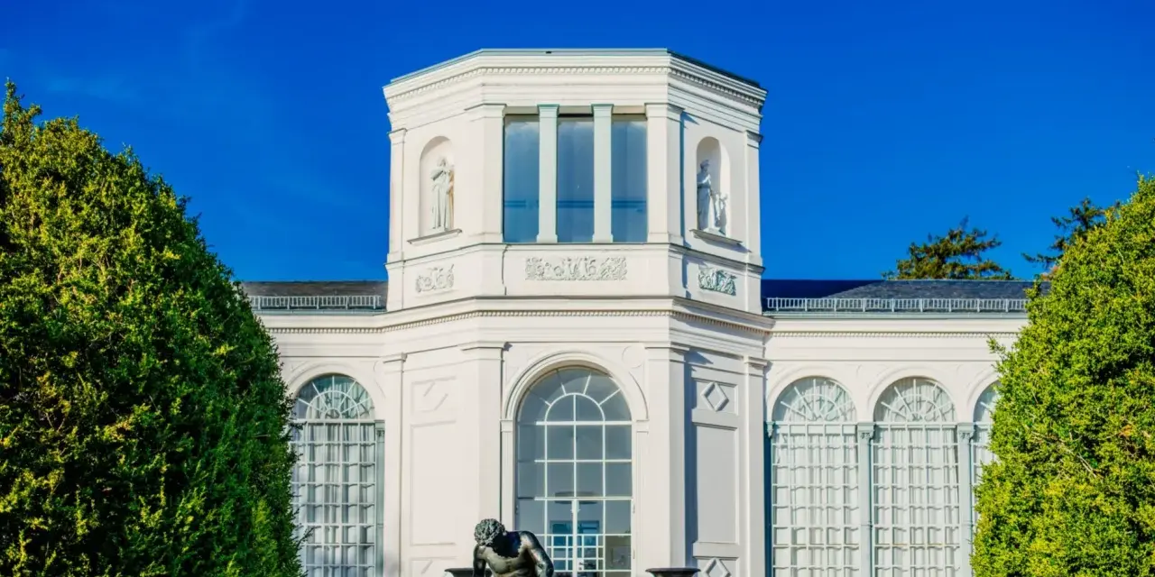 Die Orangerie Putbus, ein strahlend weißes Gebäude mit Skulpturen und großen Fenstern, wird von üppigem Grün eingerahmt.