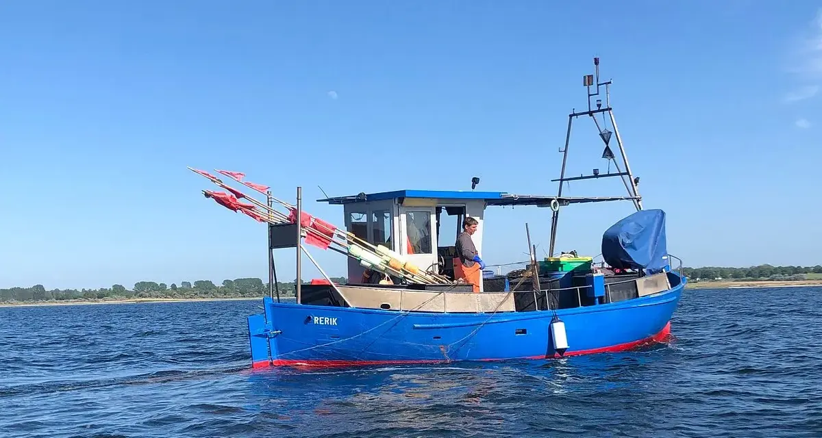 Blaues Fischerboot "RERIK" mit roten Markierungen auf dem Wasser. Ein Mann steht an Deck, rote Fahnen ragen empor.