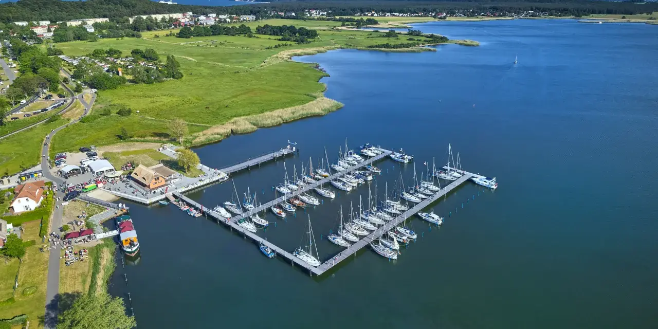 Der Hafen Sellin ist voller Segelboote, die auf dem ruhigen Wasser liegen. Im Hintergrund erstreckt sich eine grüne Landschaft mit Häusern und Bäumen.