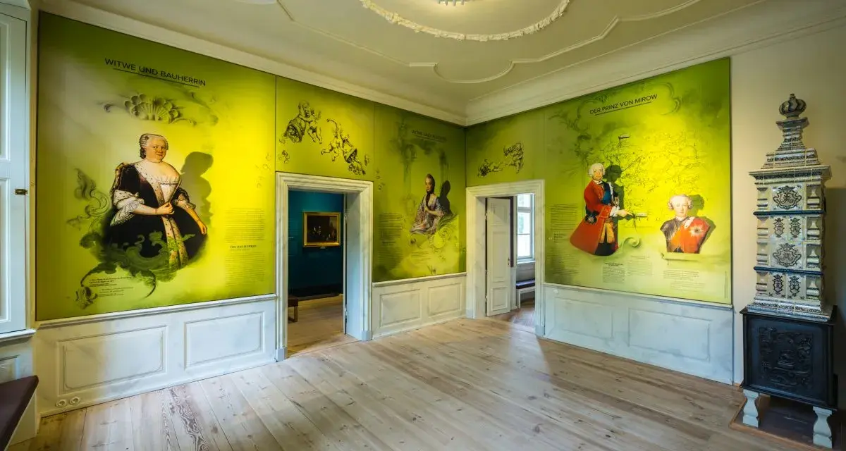 Ein Raum im Schloss Mirow mit Wandmalereien, die historische Persönlichkeiten und Karten zeigen. Ein Kachelofen steht rechts.