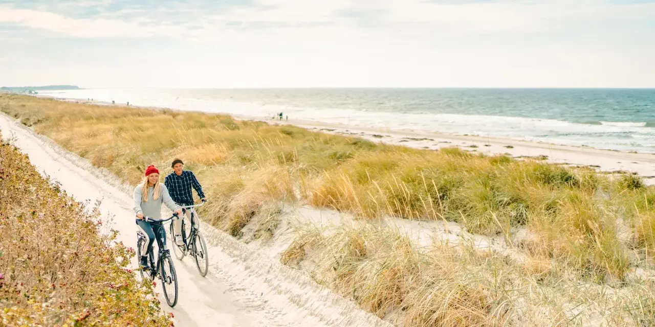 Ein Paar genießt seine baltischen Abenteuer auf Fahrrädern entlang eines sandigen Weges am Meer, umgeben von Dünen und Gräsern.