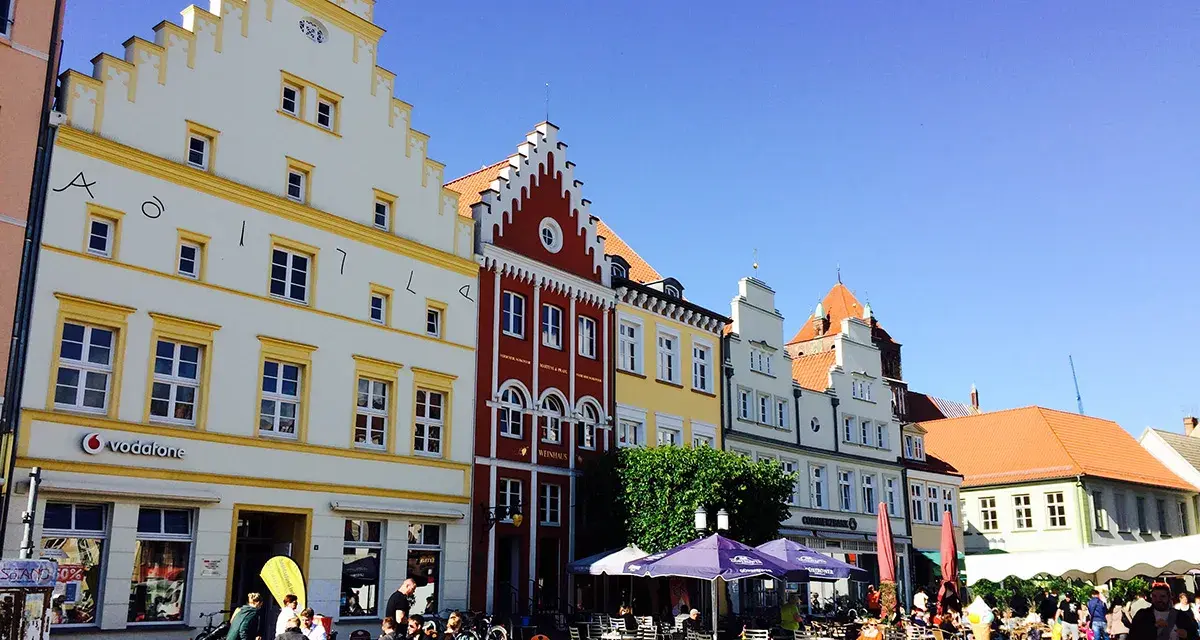 Bunte Häuserfronten säumen den mv markt. Menschen sitzen in Cafés unter Sonnenschirmen und genießen den sonnigen Tag.