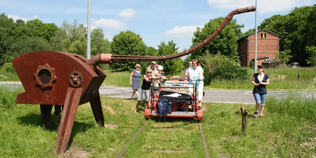 Eine Gruppe von Menschen auf einer Draisine Dargun, neben einer rostigen Skulptur.