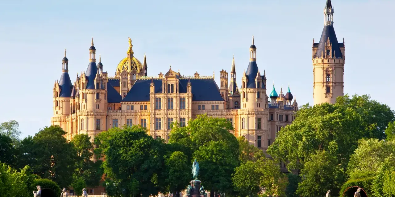 Das Schweriner Schloss, ein Märchenschloss mit goldenen Kuppeln und Türmen, thront majestätisch über einem grünen Park mit Statuen und einem Teich.